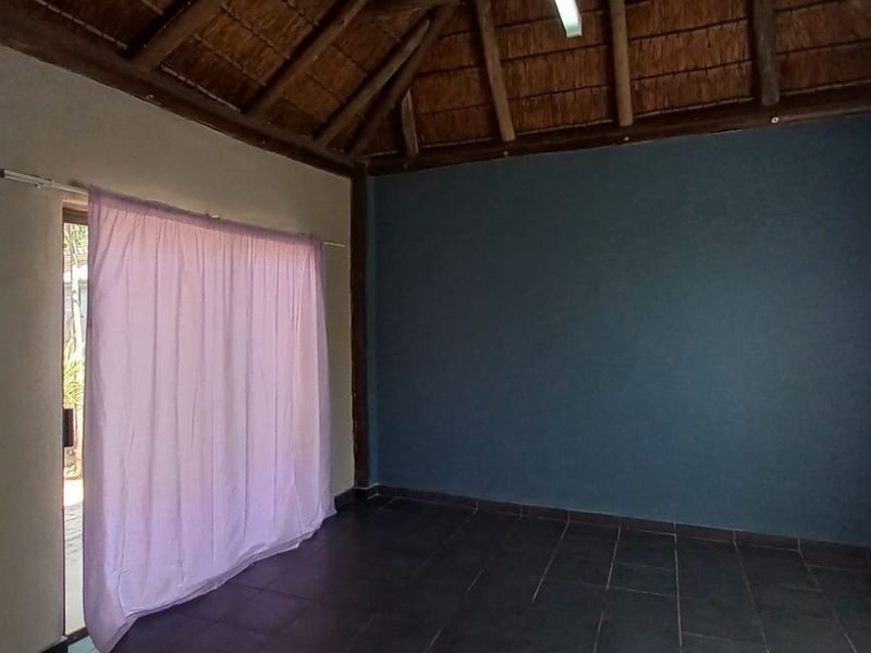An empty living room featuring a high thatched ceiling with exposed timber beams, dark tiled flooring, and a sliding door leading to the exterior.