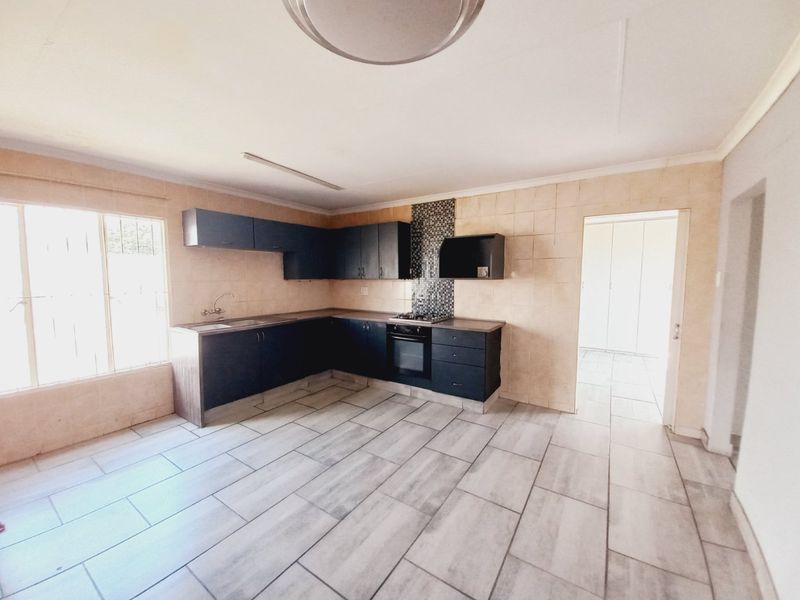 A functional kitchen featuring dark cabinetry, integrated oven and hob, tiled splashback, and neutral floor tiles in an open-plan layout.