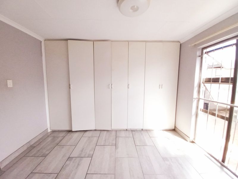A bright, empty bedroom featuring neutral tiled flooring and extensive built-in cupboard storage with access to an exterior area via a security gate.