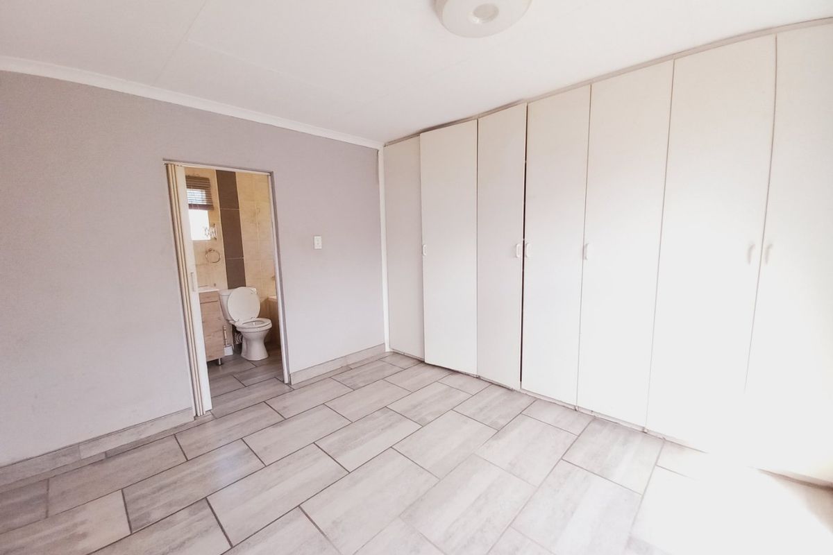 A bright, empty bedroom featuring neutral tiled flooring and extensive built-in cupboard storage, with an adjoining en-suite bathroom visible through the doorway.