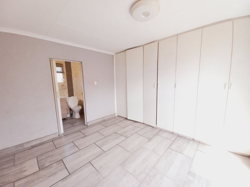 A bright, empty bedroom featuring neutral tiled flooring and extensive built-in cupboard storage, with an adjoining en-suite bathroom visible through the doorway.