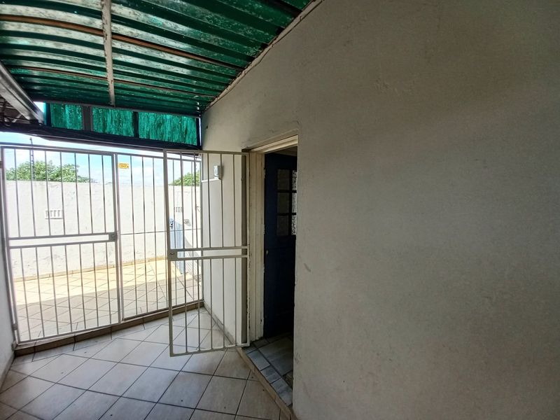 A covered outdoor patio area featuring tiled flooring, a security gate, and a corrugated roof structure, providing a functional transition space.
