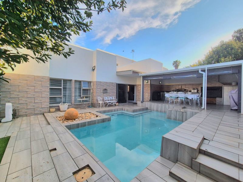 A well-maintained outdoor entertainment area featuring a swimming pool, tiled decking, and a covered patio with a built-in braai.