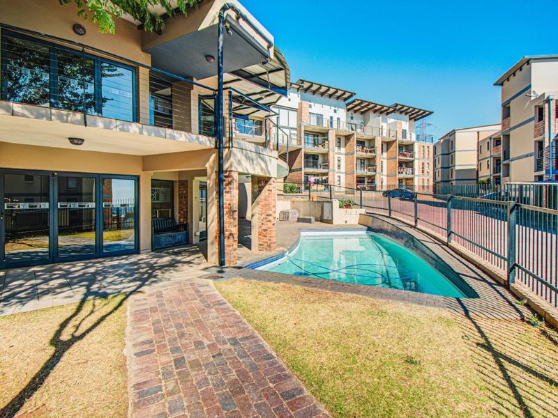 A communal pool area featuring brick paving, metal fencing, and access to the apartment building's covered patio and balcony levels.
