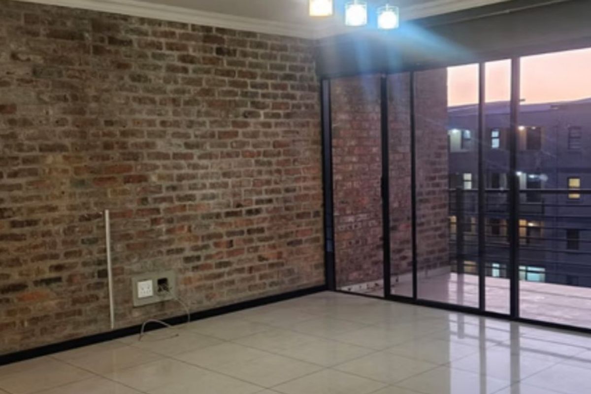 An empty living room featuring an exposed brick feature wall, large sliding glass doors leading to a balcony, and modern tiled flooring.