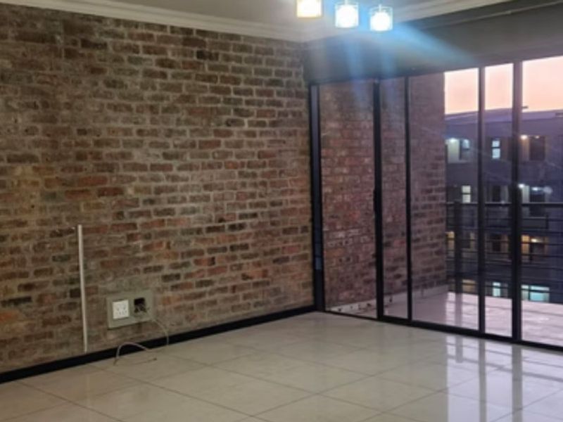 An empty living room featuring an exposed brick feature wall, large sliding glass doors leading to a balcony, and modern tiled flooring.
