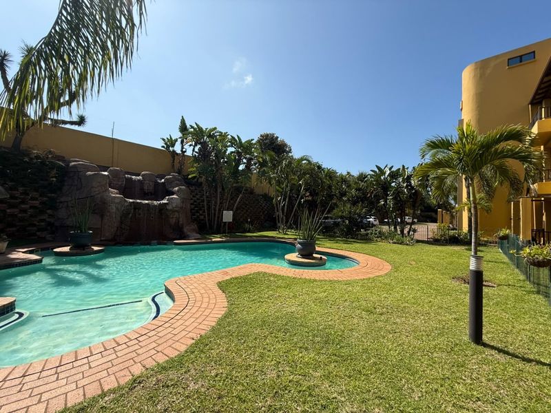 A well-maintained communal pool area featuring a decorative rock waterfall, brick paving, and lush landscaped garden surroundings within an apartment complex.