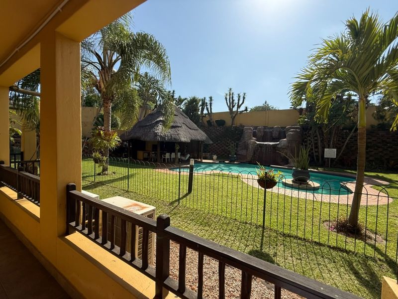 Outdoor pool area featuring a swimming pool, rock waterfall feature, and a thatched lapa set within a landscaped garden with perimeter fencing.