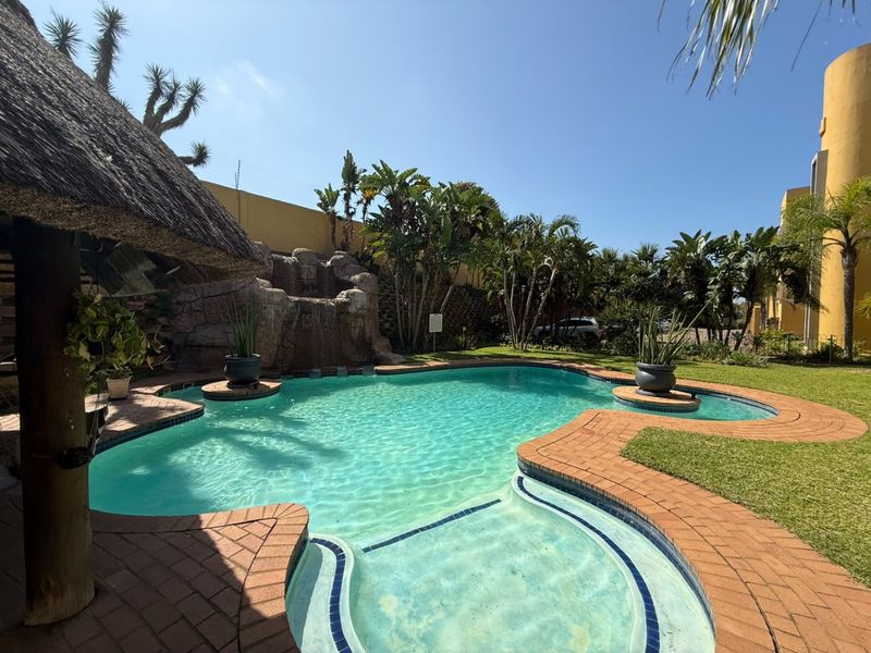 A well-maintained outdoor pool area featuring a rock waterfall, brick paving, and a thatched lapa, set within a landscaped garden environment.