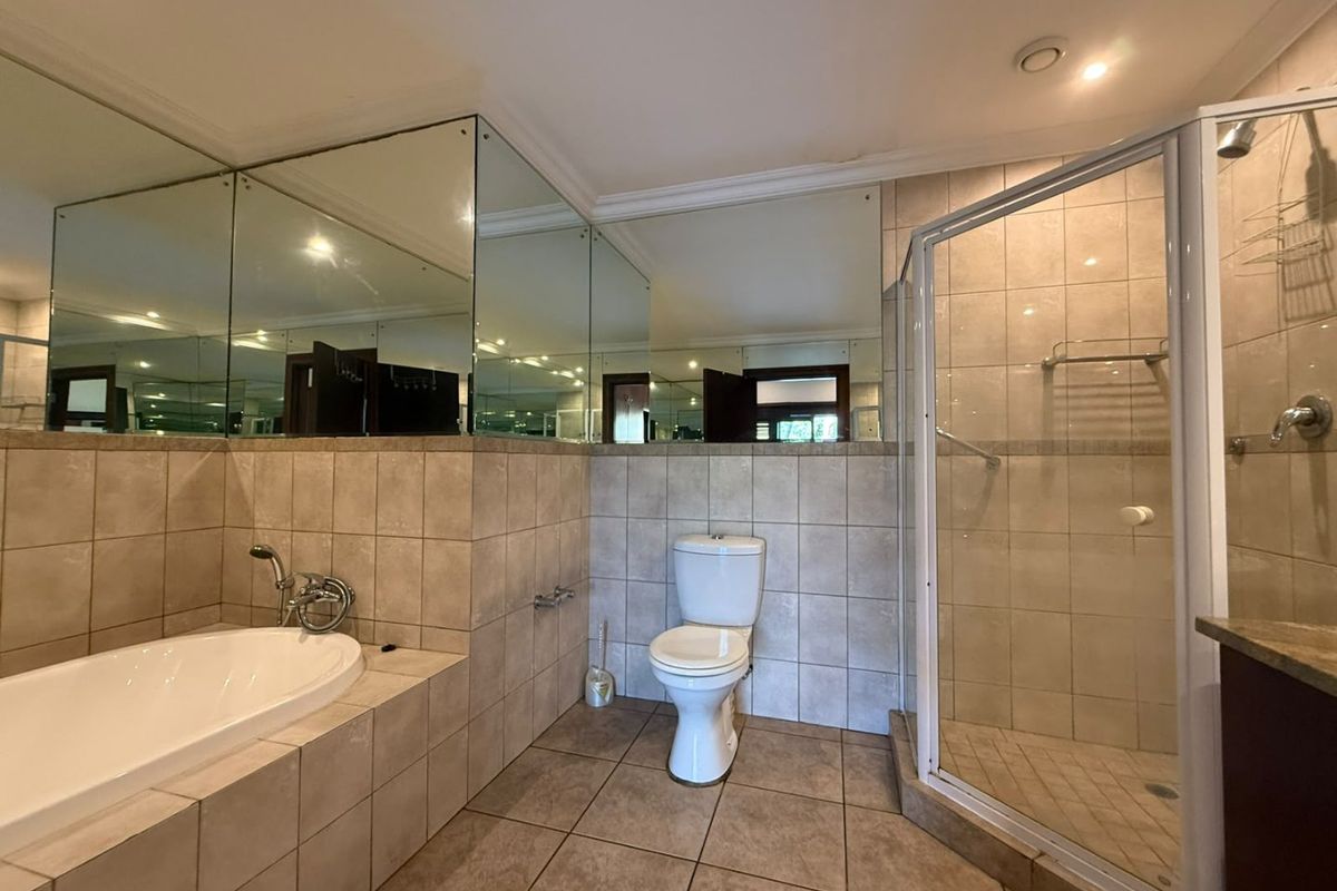A spacious bathroom featuring a built-in bathtub, a corner shower enclosure, a toilet, and extensive wall-mounted mirrors with neutral floor and wall tiling.