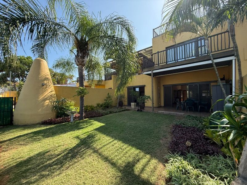 Rear view of the apartment featuring a landscaped garden, a covered patio area, and an upper-level balcony with railings.