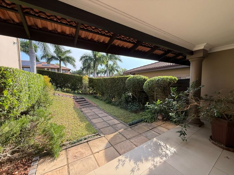A covered patio area featuring tiled flooring and a support column, overlooking a well-maintained garden with a paved pathway and established hedge boundaries.
