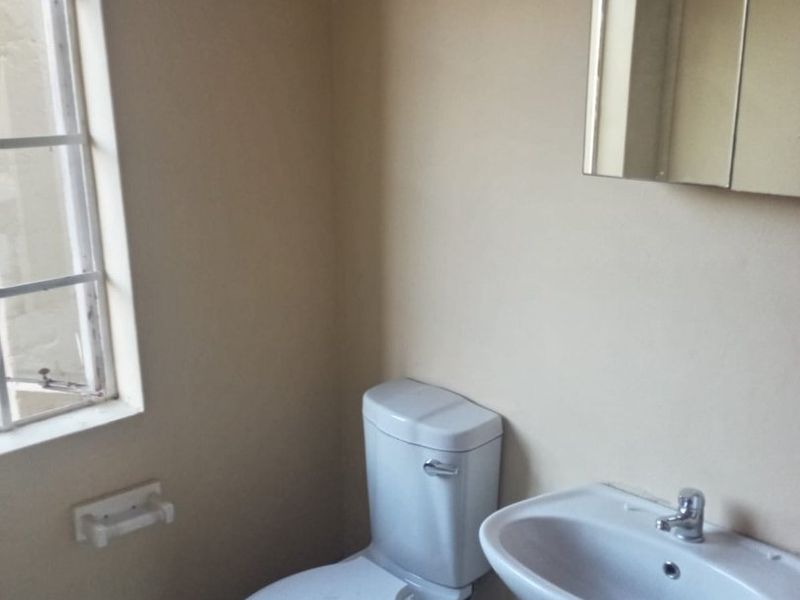 A functional bathroom featuring a pedestal basin, toilet, and wall-mounted mirror cabinet, finished with tiled flooring and a window with security bars.