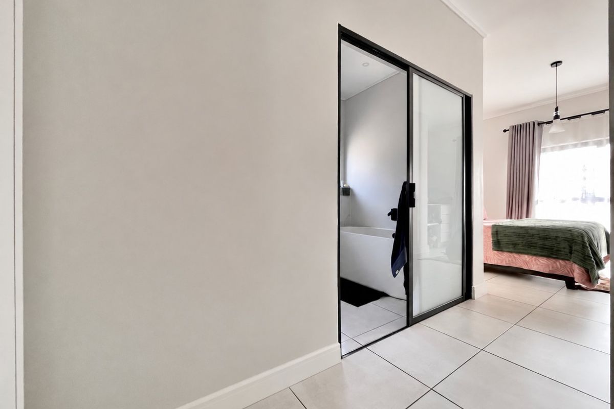 A modern bedroom featuring tiled flooring, clean neutral walls, and a sleek black-framed glass door leading to an en-suite bathroom.