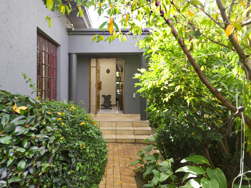 A view of a rear entrance featuring a brick paved pathway leading to a covered patio area with tiled steps and established garden surroundings.