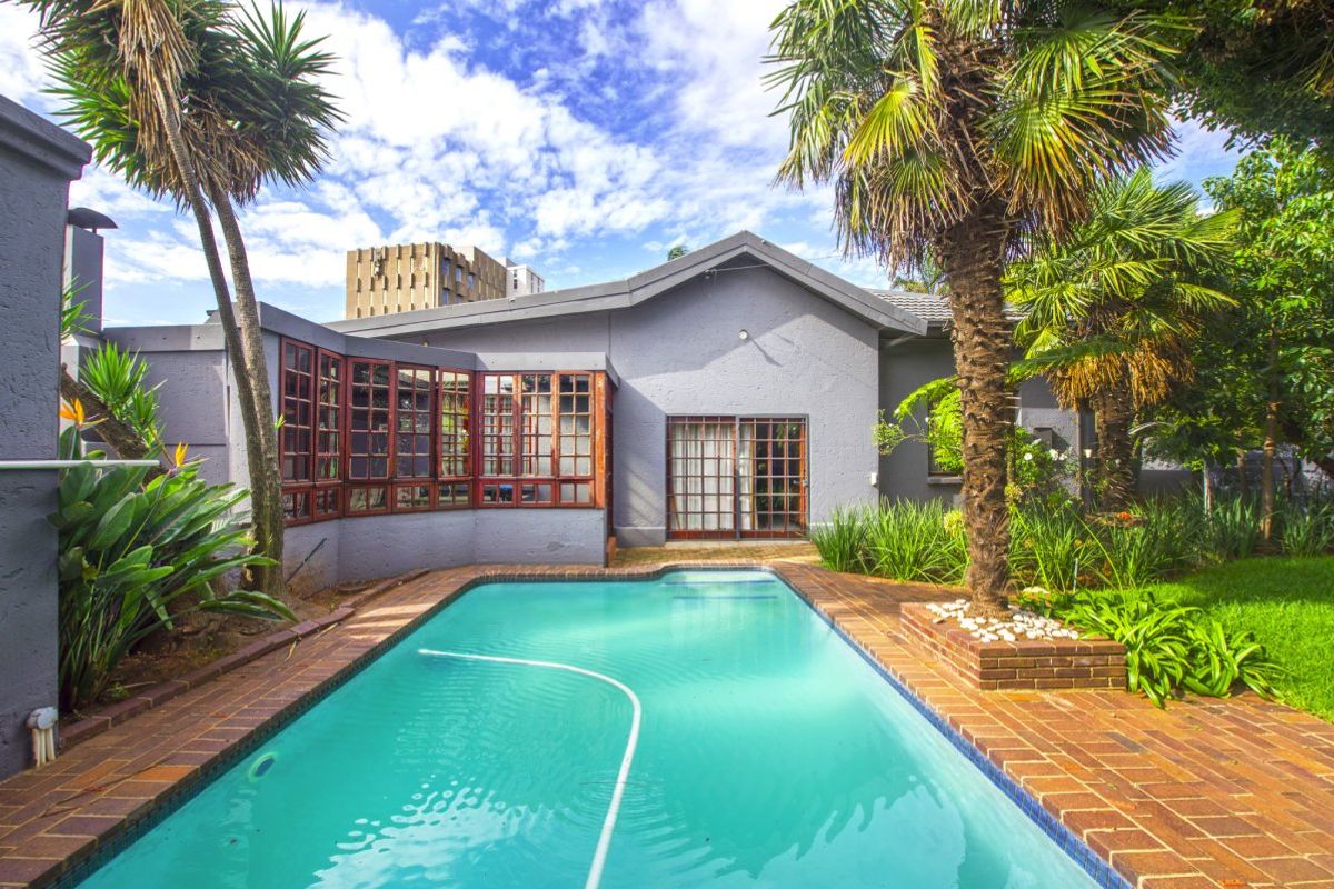 A well-maintained backyard featuring a swimming pool with brick coping, surrounded by established palm trees and a landscaped garden area.