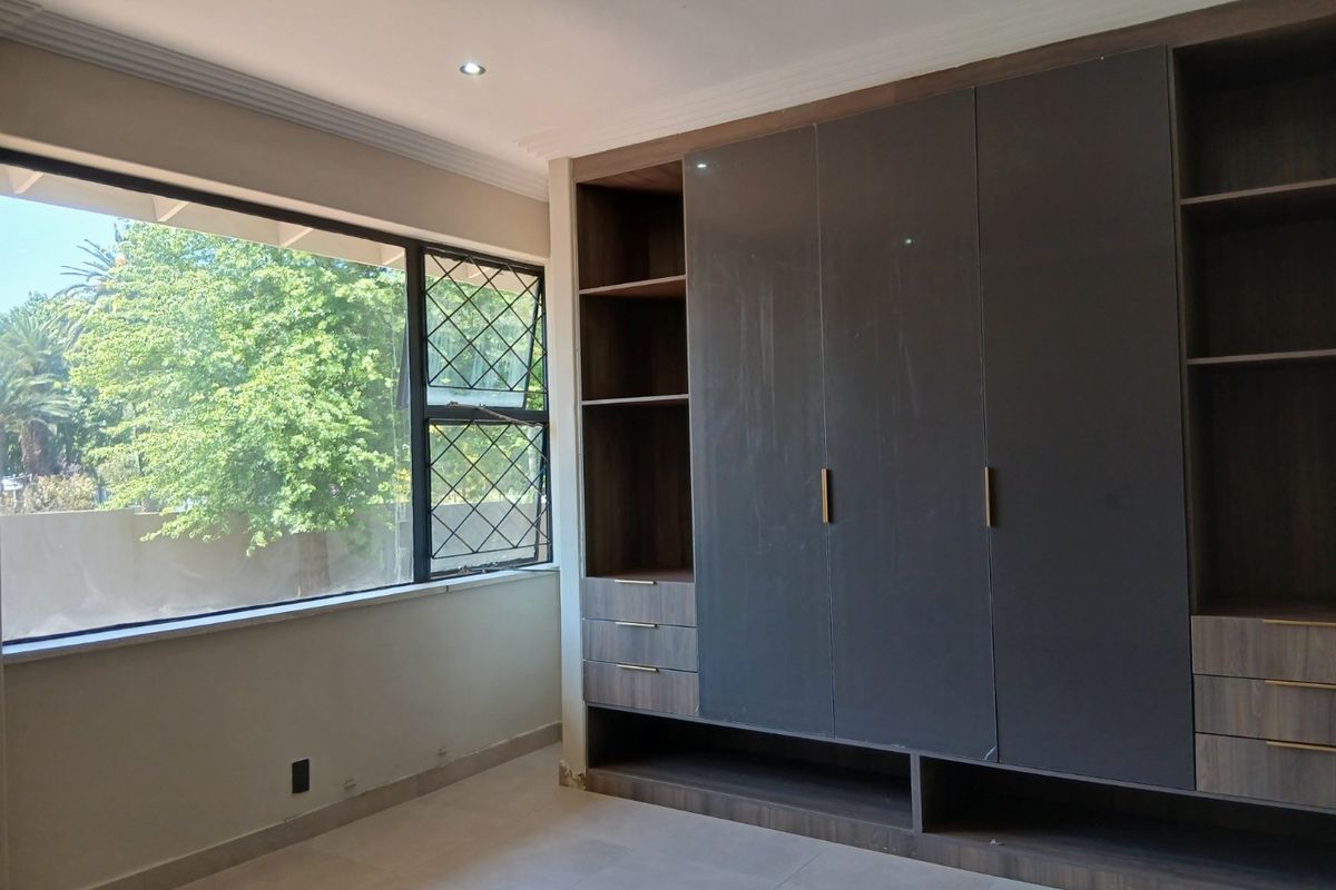 A bright, empty bedroom featuring modern dark grey built-in cupboards, tiled flooring, and large windows with security bars.