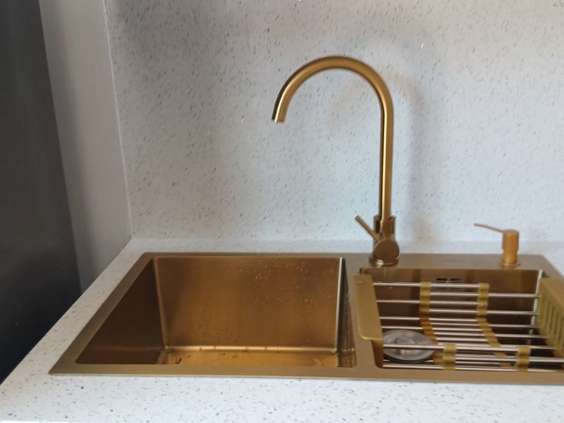 A modern kitchen sink area featuring a gold-toned stainless steel sink, matching gooseneck faucet, and a clean, speckled stone-look countertop and splashback.