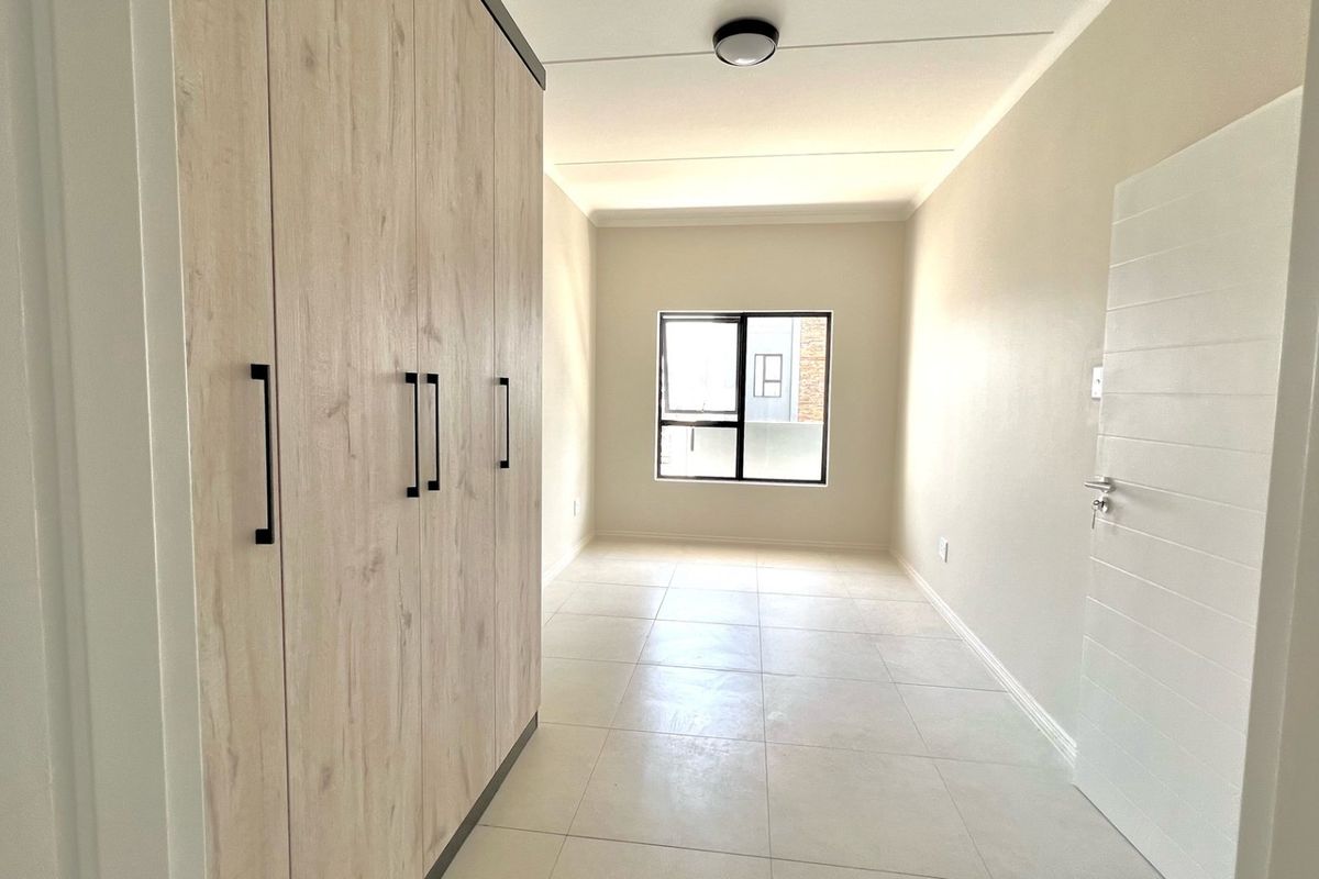 A bright, empty bedroom featuring modern light wood built-in cupboards, neutral tiled flooring, and a large aluminium-framed window.