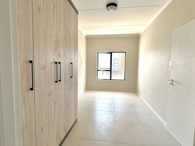 A bright, empty bedroom featuring modern light wood built-in cupboards, neutral tiled flooring, and a large aluminium-framed window.