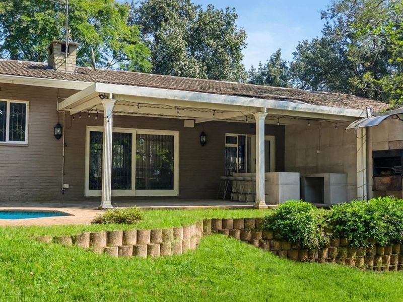 Rear view of the house featuring a covered patio with a built-in braai, swimming pool, and landscaped garden with a retaining wall.