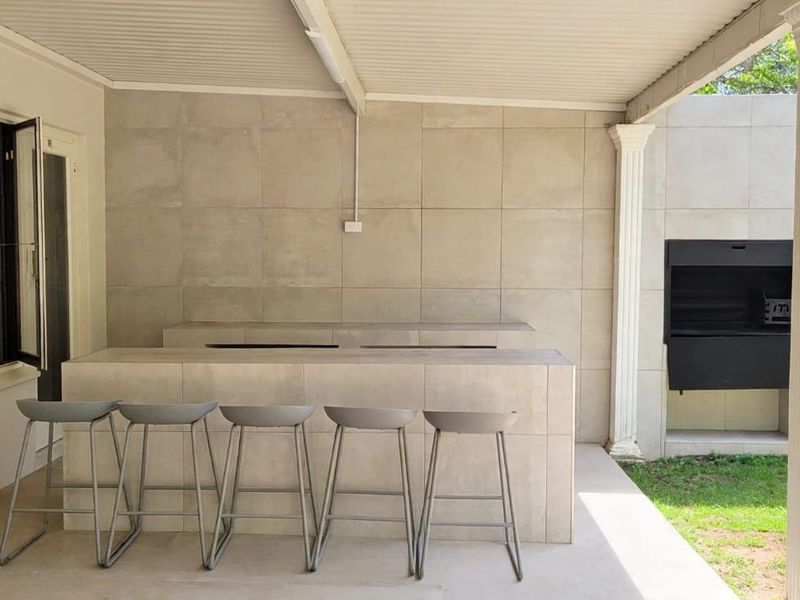 Covered outdoor patio featuring a built-in braai, tiled bar counter, and tiled wall finishes, overlooking a garden area.