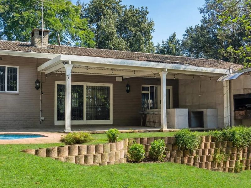 Rear view of the property featuring a covered patio with a built-in braai, overlooking a swimming pool and landscaped garden with retaining walls.