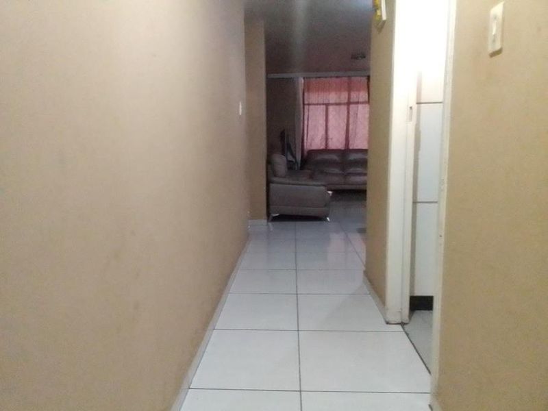 A narrow interior hallway featuring tiled flooring and plain wall finishes, leading towards an adjacent living area.