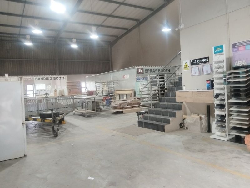 Spacious industrial workshop area featuring concrete flooring, high-clearance metal roof structure, dedicated spray and sanding booths, and an integrated office staircase.