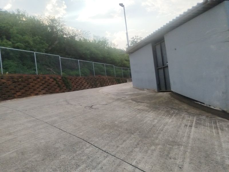Exterior rear view of the industrial property featuring a concrete driveway, retaining wall, and perimeter wire mesh fencing.