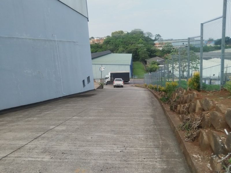 A concrete driveway leading to the rear of an industrial property, featuring security fencing and a retaining wall along the perimeter.