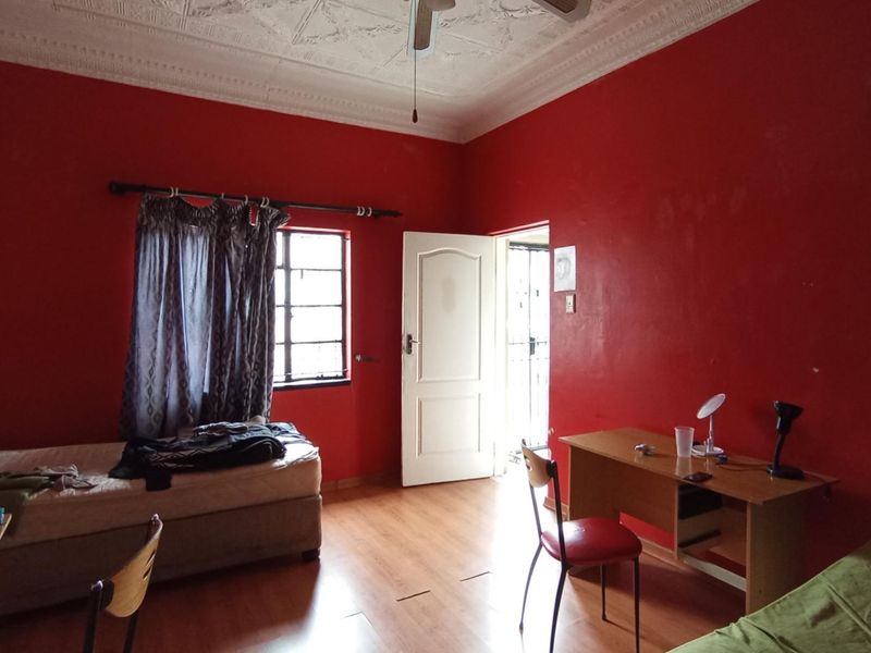 A bedroom featuring laminate flooring, an ornate ceiling with a fan, and a window, currently used as a study and sleeping area.