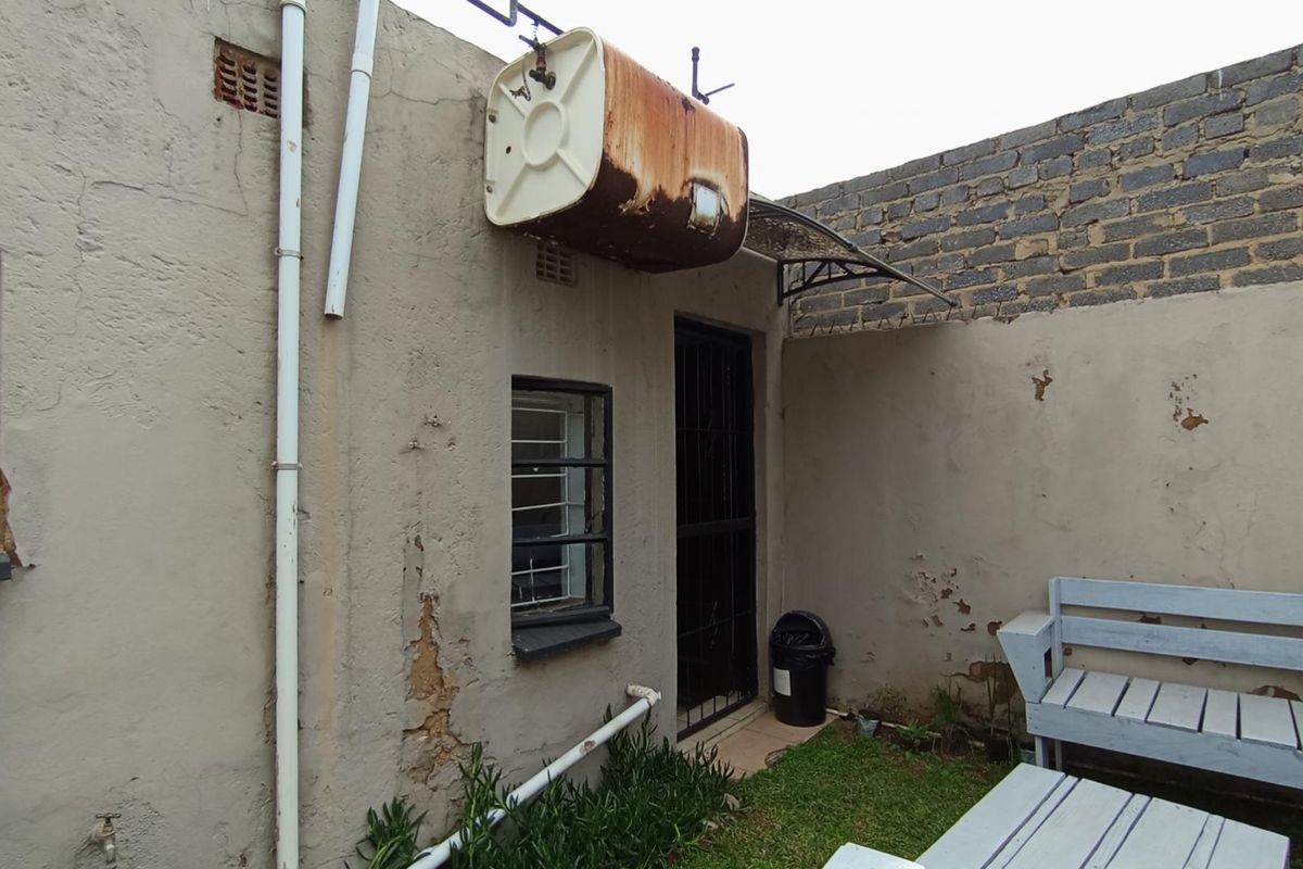 Rear exterior view showing a weathered wall with a rusted external geyser, security features, and signs of significant plaster and paint deterioration.