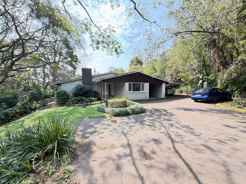 Front exterior view of a single-story house featuring a paved driveway, carport, brick chimney, and established garden surroundings.