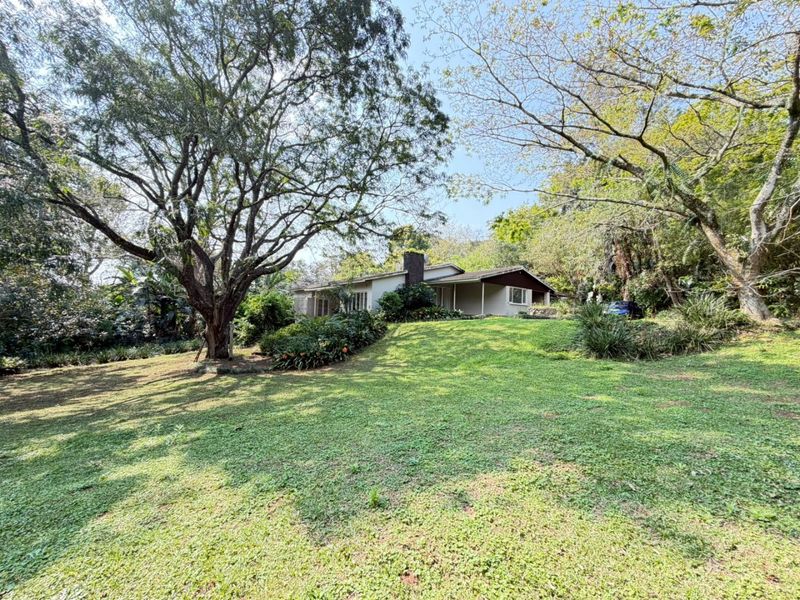 A spacious rear garden featuring mature trees, a lawn, and a view of the house with a brick chimney and covered patio area.