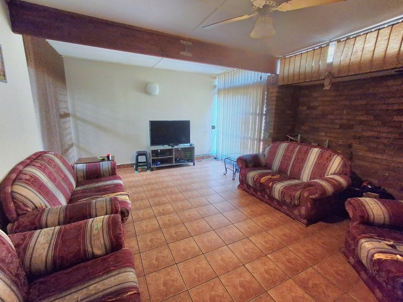 Spacious living room featuring a rustic brick accent wall, exposed timber ceiling beam, ceiling fan, and tiled flooring throughout.