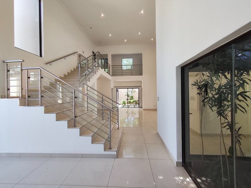 Spacious double-volume entrance hall featuring a modern staircase with stainless steel and glass balustrades, polished porcelain tiles, and abundant natural light.