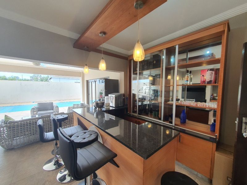 A well-appointed indoor bar area featuring a granite-topped counter, custom wooden cabinetry with mirrored shelving, and elegant pendant lighting, overlooking the outdoor pool area.