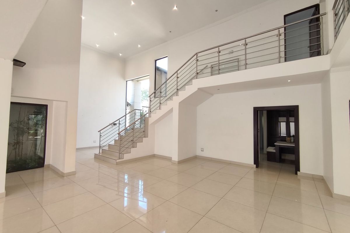 Spacious double-volume living area featuring polished tile flooring, a modern staircase with stainless steel balustrades, and recessed ceiling lighting.