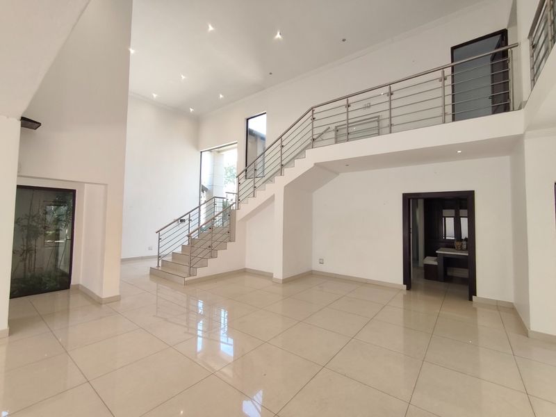Spacious double-volume living area featuring polished tile flooring, a modern staircase with stainless steel balustrades, and recessed ceiling lighting.