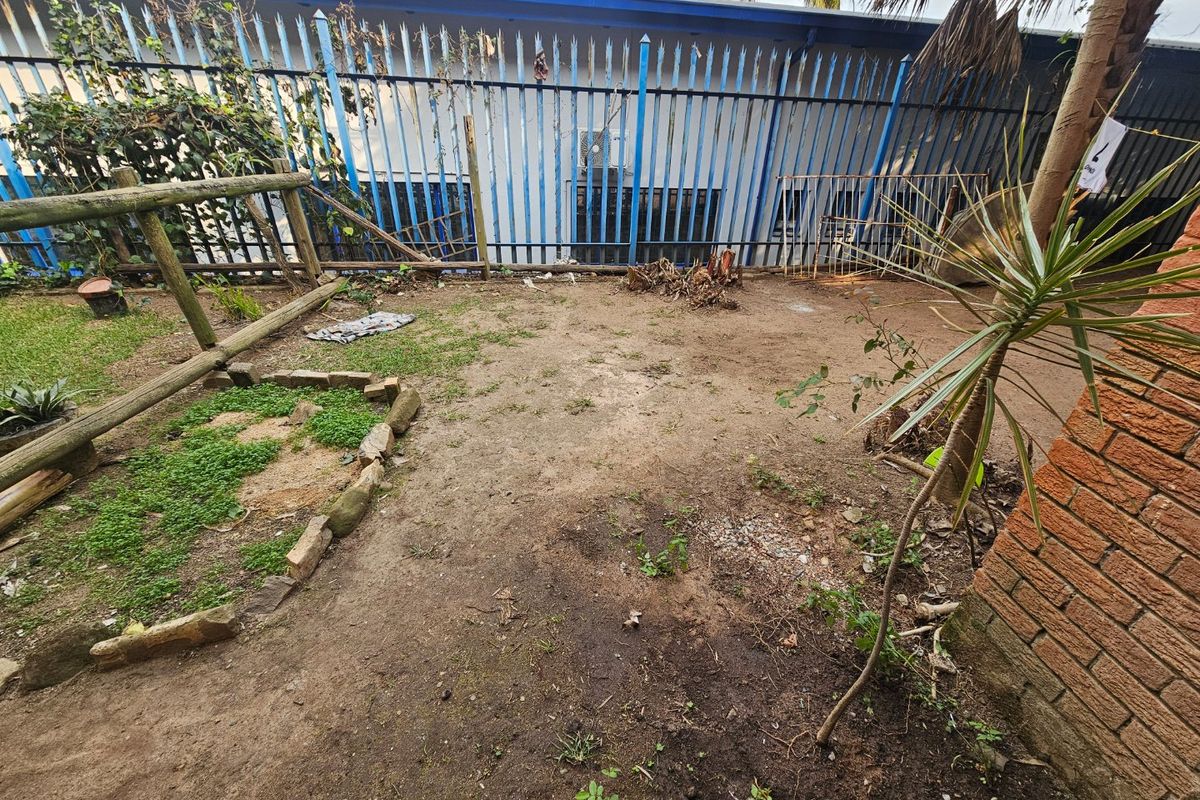A small, unlandscaped garden area featuring a metal perimeter fence, brick walling, and a timber log border, requiring significant maintenance and landscaping.