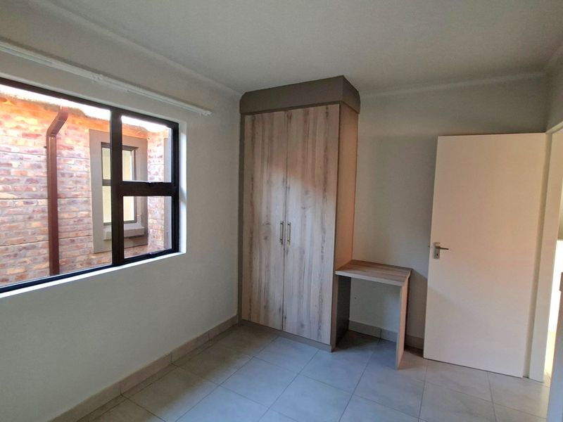 A clean, empty bedroom featuring neutral tiled flooring, a built-in wardrobe with an integrated desk, and a large window overlooking a brick courtyard.