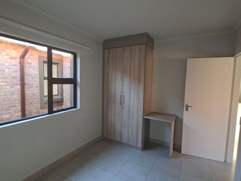 A clean, empty bedroom featuring neutral tiled flooring, a modern built-in wardrobe with an integrated desk, and a window overlooking a brick courtyard.