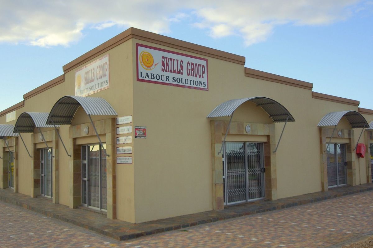 A commercial corner building featuring a beige facade, stone accents, corrugated metal awnings over multiple entrances, and paved exterior grounds.
