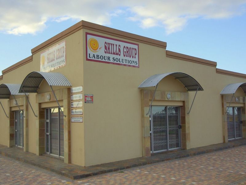 A commercial corner building featuring a beige facade, stone accents, corrugated metal awnings over multiple entrances, and paved exterior grounds.