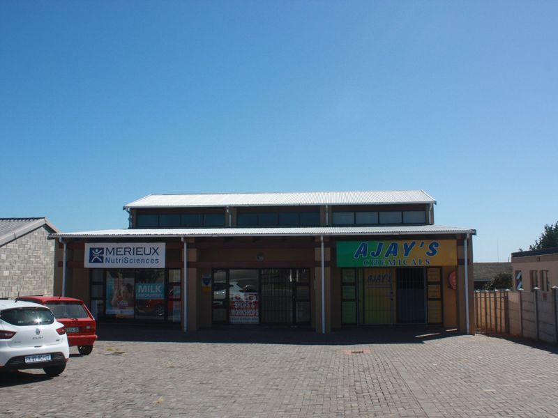 Front elevation of a commercial property featuring a paved parking area, corrugated metal roof, and multiple storefront units with glass facades.