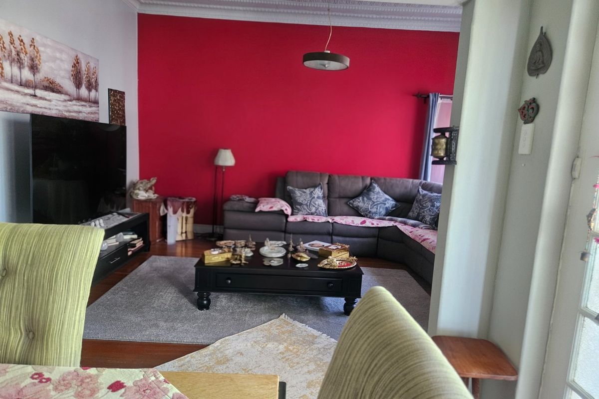 A living room featuring hardwood flooring, decorative ceiling cornicing, and a bold red accent wall, viewed from an adjacent dining area.