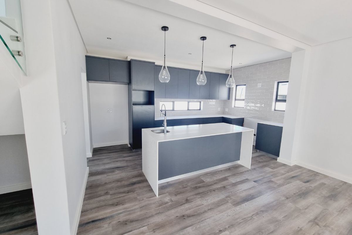 Modern, empty kitchen featuring a central island, dark cabinetry, subway tile backsplash, and wood-look laminate flooring in a bright, open-plan layout.