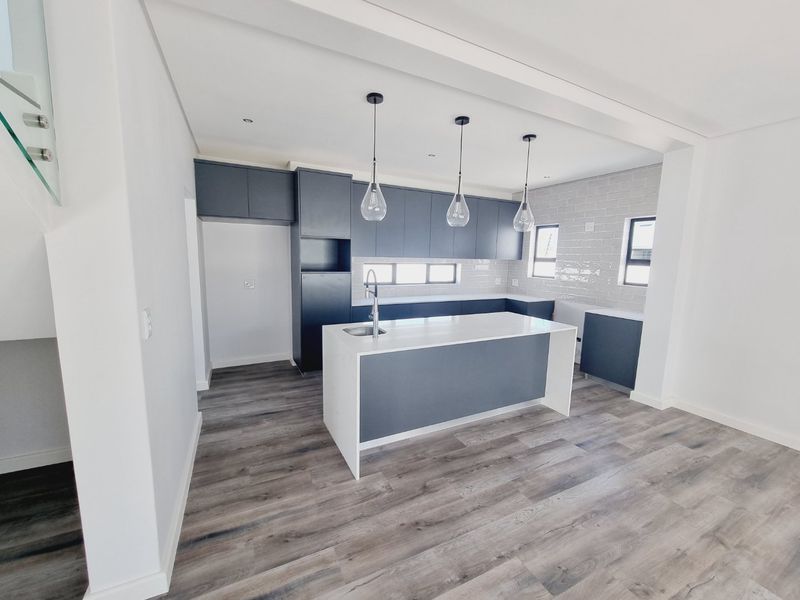 Modern, empty kitchen featuring a central island, dark cabinetry, subway tile backsplash, and wood-look laminate flooring in a bright, open-plan layout.