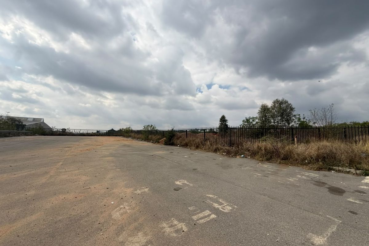 A vacant residential land plot featuring an asphalt surface and metal perimeter fencing under a cloudy sky.